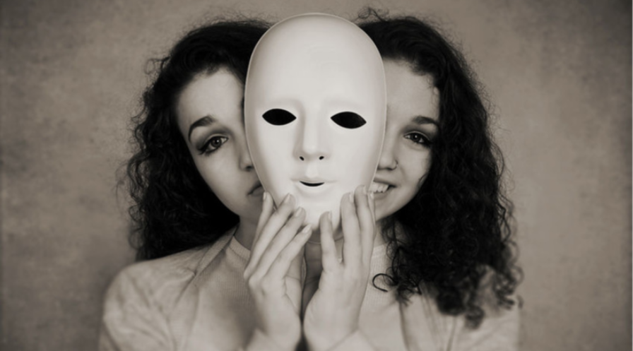 Zdjęcie czarno białe dwie te same kobiety tak jakby połączone stoją obok siebie. Kobieta z lewej ma smutną twarz, kobieta z prawej uśmiechnięta. Na wysokości głowy pomiędzy twarzami trzymają w ręku białą maskę.