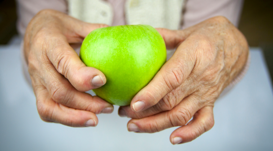 Zielone jabłko trzymane w dłoniach seniora z powykrzywianymi palcami.